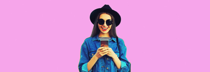 Portrait of stylish happy smiling young woman model with smartphone wearing black round hat, jean jacket on pink background, blank copy space for advertising text