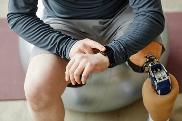 Close up of man with prosthetic leg checking smartwatch sitting on fitness ball during home workout