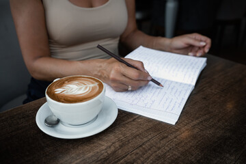 Unrecognizable woman sits at table in cafe with cup of coffee and writes in notebook notes writing information
