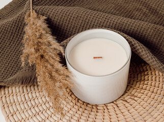 Candle in white glass jar on wicker mat with pampas grass and brown towel at the background, home...