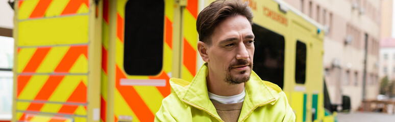 Paramedic in jacket looking away near blurred ambulance car on street, banner. © LIGHTFIELD STUDIOS
