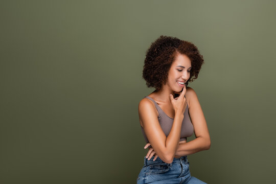 Positive african american woman in top touching chin isolated on green.