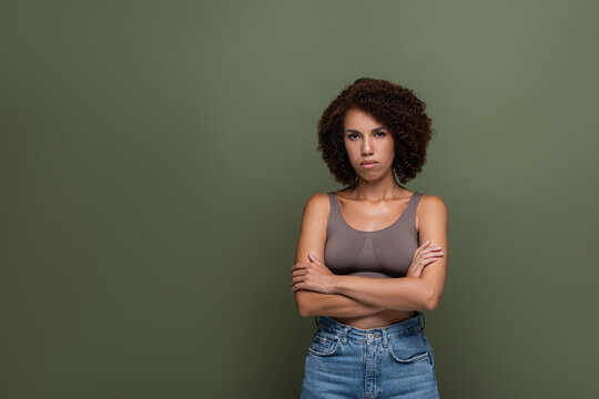 Serious african american woman in top and jeans crossing arms isolated on green.