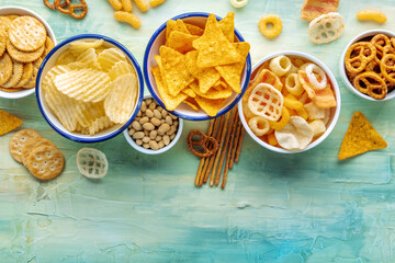 Potato and tortilla chips and other salty snacks, shot from above with copy space. Party food on a teal blue background. A mix of appetizers in bowls