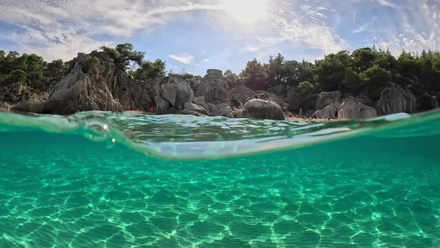 Half underwater slow-motion shot of tropical beach with crystal clear sea water. View from the sea. Sandy beach, rocks, trees and water surface