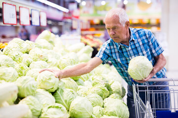 Mature senor examines cole in the vegetables section of supermarket - obrazy, fototapety, plakaty