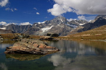 Landschap met een glad oppervlak van het meer Riffelsee, bergen en wolken erin weerspiegeld, met een grote steen op de voorgrond, op een berg Gornergrat, nabij Zermatt, in Zuid-Zwitserland