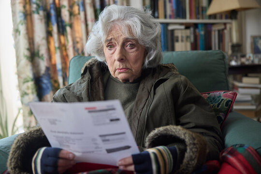 Senior Woman Wearing Coat Indoors Trying To Keep Warm At Home In Energy Crisis Looking At Energy Bill