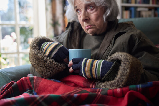 Senior Woman Wearing Coat Indoors With Hot Drink Trying To Keep Warm At Home In Energy Crisis