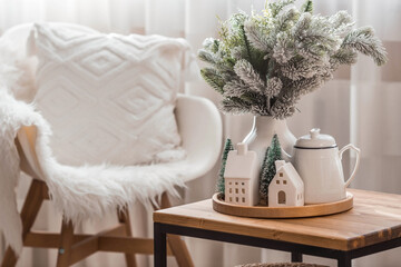 A bouquet of fir trees, a plaid in a wicker basket and Scandinavian white houses on a wooden table in the home interior of the living room. A cozy concept of festive home decoration.