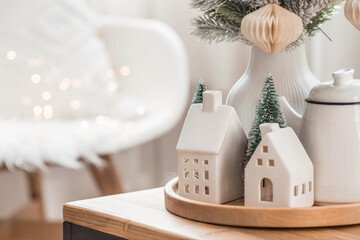 A bouquet of fir trees, a plaid in a wicker basket and Scandinavian white houses on a wooden table in the home interior of the living room. A cozy concept of festive home decoration.