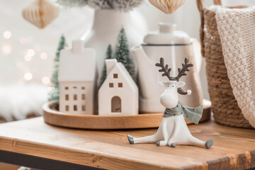 Fototapeta na wymiar A bouquet of Christmas trees, reindeer, a blanket in a basket and Scandinavian white houses on a wooden table in the home interior of the living room. Cozy concept of festive decoration of the house.