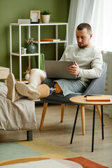 Vertical full length portrait of adult man with prosthetic leg relaxing at home and using laptop