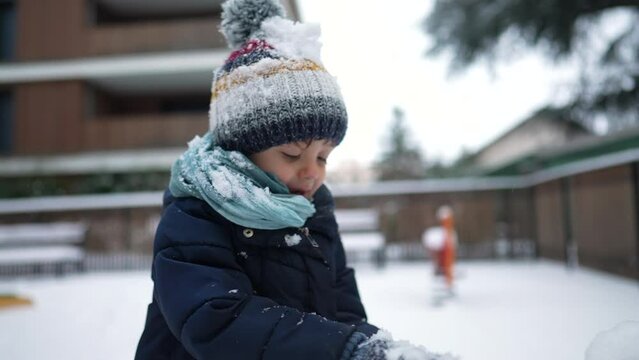 Small boy playing with snow outside in winter season. Parent throwing snowball at child wearing beanie and coat in slow motion