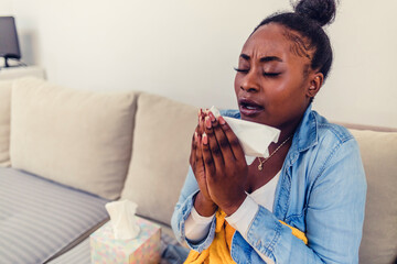 Shot of a young woman blowing her nose with a tissue at home. Young woman with a cold, sitting on...