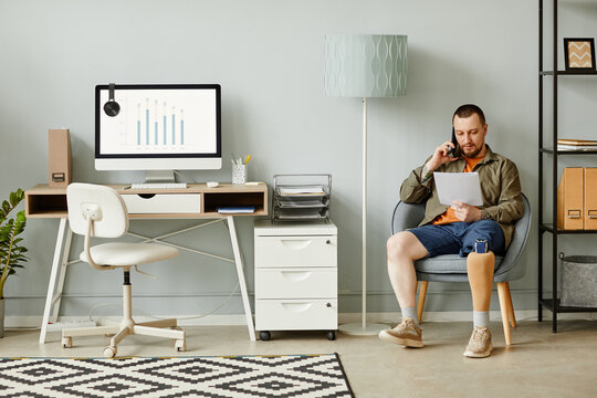 Full length portrait of man with prosthetic leg calling by phone while sitting in chair in home office interior, copy space