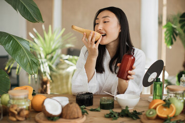 Beautiful asian woman preparing body milk or cream from pumpkin for skin care sitting at wooden table with various ingredients for making natural cosmetics on light exotic studio background.