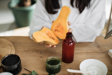 Cropped view of beautiful healthy asian woman in white bathrobe sitting at wooden table with various ingredients preparing natural cosmetics at home, holding pumpkin and bottle with body milk or cream