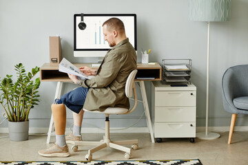 Minimal full length portrait of man with prosthetic leg working at home office and reading...