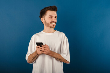 Cheerful young man wearing t-shirt posing isolated over blue background dreamy look, thinking while holding smartphone and looking aside.