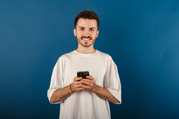 Happy young man wearing white t-shirt posing isolated over blue background holding smart phone and looking at the camera with big smiles. Messaging, watching or scrolling on social media.