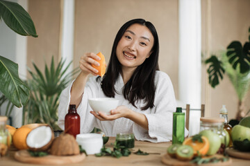 Beautiful asian woman preparing cream using orange for skin care sitting at wooden table with various ingredients for making natural cosmetics on light exotic studio background.