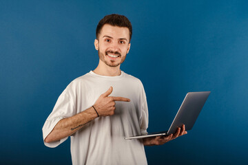 Young man smiling confident wearing white tee posing isolated over blue background smiling and pointing with index finger to the laptop pc computer.