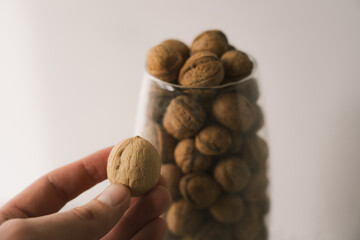 A walnut in a shell in a hand next to a glass vase with walnuts and a white blurred background.