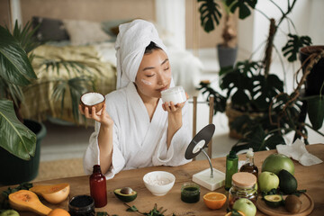 Young asian woman in white bathrobe and towel holding half of coconut and making homemade cream for healthy skin. A lot of ingredients for homemade cosmetics lying on wooden table.