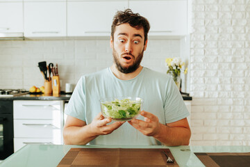  man grimaces very expressively at the sight of a salad of greens, holding a bowl in his hands. He...