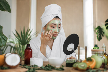 Attractive asian woman looking at mirror sitting at table with ingredients for homemade cosmetics...