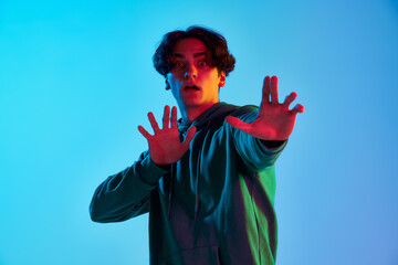 Portrait of young man with curly hair posing with hands forward, expressing fear isolated over blue background in neon light