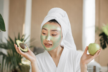 Portrait of beautiful young asian woman with green cosmetic mask apple and avocado in her hands...