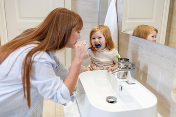 Mother watching daughter brushing teeth at home. Happy woman brushing teeth with her daughter. Little girl brushing teeth with her mom. Smiling mother and daughter brushing teeth at home