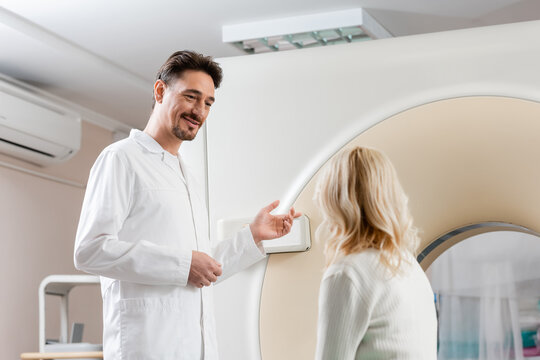 smiling doctor pointing at computed tomography scanner near blonde middle aged woman.