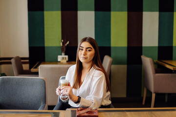 Portrait of beautiful girl, drinking hot tea or coffee in a cafe with her cell mobile phone.