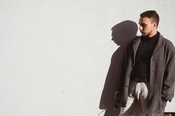 Young handsome man in dark coat and white trousers posing on sunlight against white wall, sunglasses in hand, thoughtfully looking down