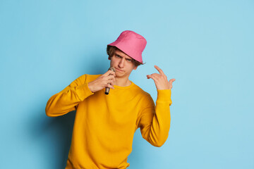 Portrait of young man with curly hair posing in pink panama and singing isolated over blue background. Famous performer