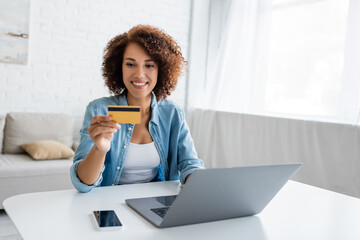 Positive african american woman holding credit card near devices at home.