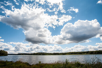 Obraz premium Picturesque landscape with a cloudy sky and a wide river. In the distance, across the river, there is a forest. An image as a background for your travel and nature illustrations.