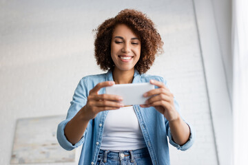 low angle view of happy african american woman with curly hair holding smartphone during video chat.