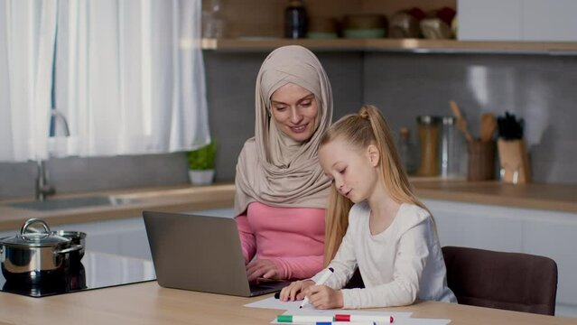 Busy muslim woman in hijab working on laptop online, little daughter drawing near at kitchen, tracking shot, free space