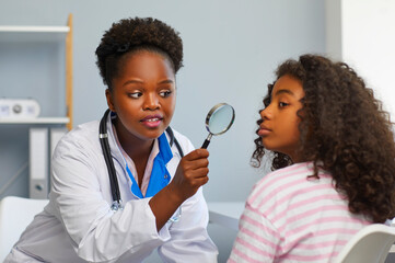 Concept of facial skin problems in adolescence. Female dermatologist examines teenage girl's face with magnifying glass for moles and redness. African American girl in dermatologist doctor's office. - obrazy, fototapety, plakaty