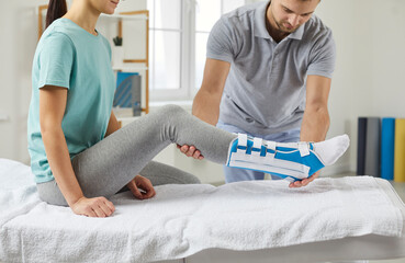 Woman with a splint on her shin lies on a couch in the physiotherapist's office in the rehabilitation center. Male physiotherapist examines a patient's leg during a rehabilitation consultation  - obrazy, fototapety, plakaty