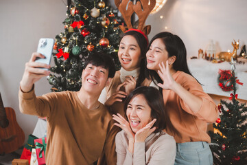 Group young Asian friends celebrating Christmas and New Year in the evening, sitting holding gift boxes taking a selfie together on the smart phone near the Christmas tree at home.