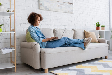 focused african american woman using laptop while sitting on couch and working from home.