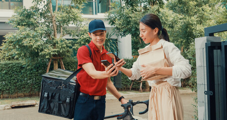 Young Asian delivery man with bicycle in red uniform carry case box knock door home online shopping paper bag to woman in front of door lady scan QR code payment on smartphone at house concept.