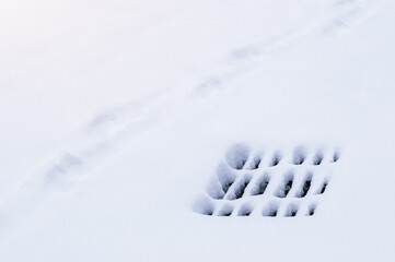 Storm sewer grate under snow during snowfall in winter. Black slits on a white smooth surface....