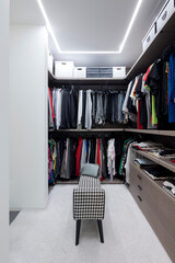 Front view walk-in closet full of clothes and in the center is a stylish stool