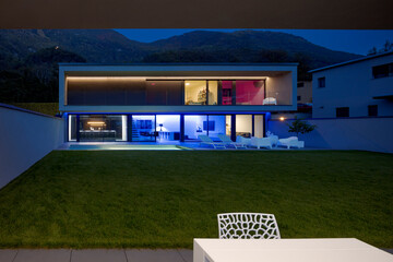 Modern house with swimming pool and garden in night scene illuminated by colored LED lights. Behind...